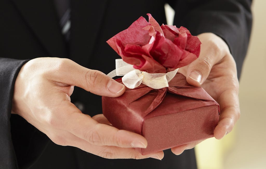 Close-up of hands presenting a beautifully wrapped gift with a red paper flower.